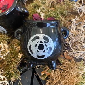 May include: Black cauldron candle holder with a white pentagram and crescent moon design. The candle holder is filled with red wax and rose petals.