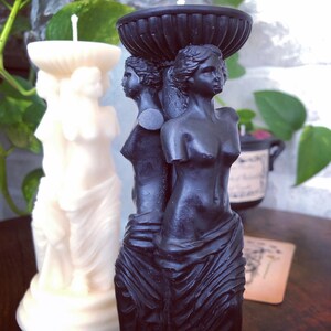 May include: Black candle holder featuring three sculpted figures of women. The figures are standing on a base with a decorative top. The candle holder is made of black wax.