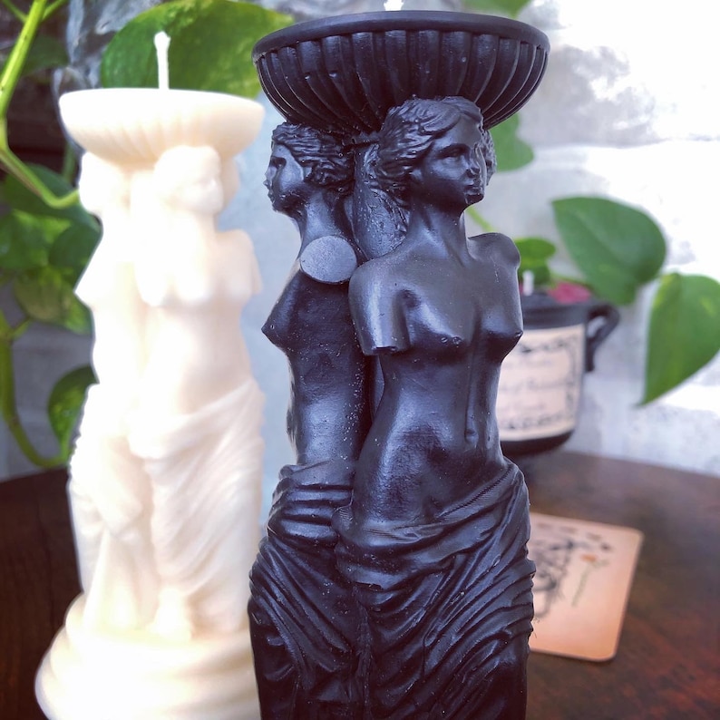 May include: Black candle holder featuring three sculpted figures of women. The figures are standing on a base with a decorative top. The candle holder is made of black wax.