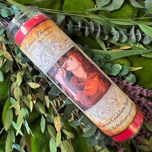 Puede incluir: Una vela cil&iacute;ndrica de cera roja en un recipiente de vidrio transparente con una etiqueta que muestra el texto Madame Phoenix Spell Candle / Bougie Magique y Warrior Goddess Spirit / Esprit de la d&eacute;esse guerri&egrave;re, representando a una persona pelirroja.