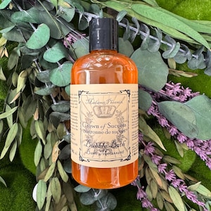 May include: An orange-colored bubble bath bottle with a black cap. The label reads "Madame Phoenix Crown of Success" and "Bubble Bath". The bottle is surrounded by green and purple foliage.