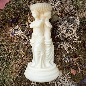 May include: White ceramic candle holder featuring three sculpted figures standing back to back. The figures are draped in flowing robes and have a classic, elegant design.