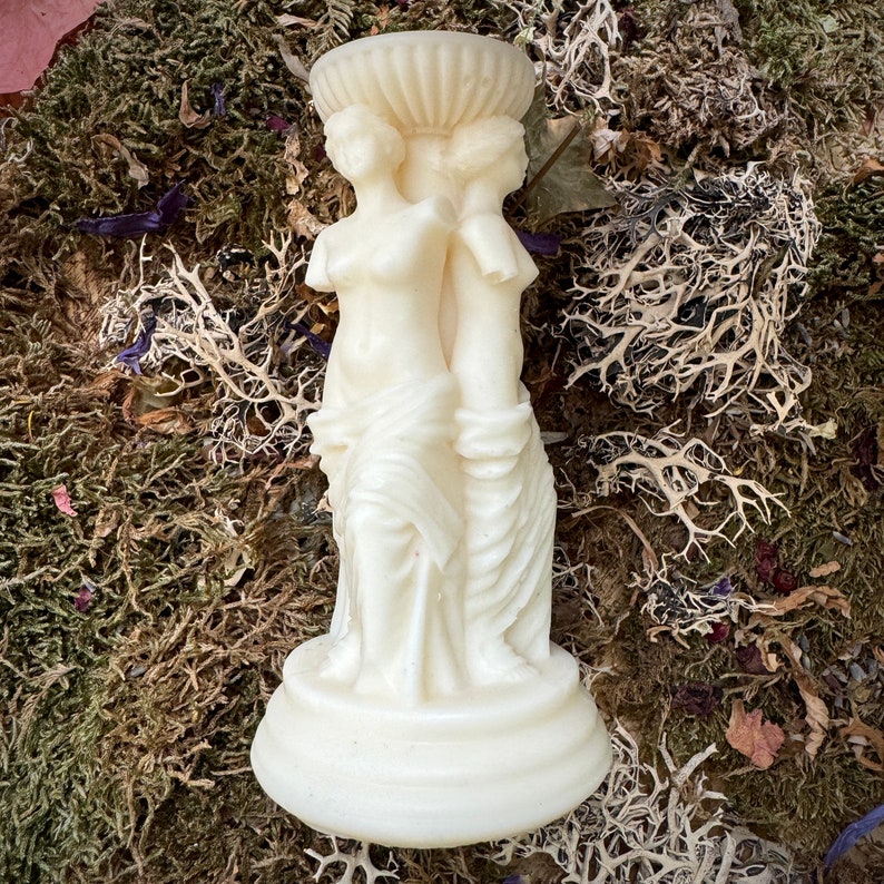 May include: White ceramic candle holder featuring three sculpted figures standing back to back. The figures are draped in flowing robes and have a classic, elegant design.