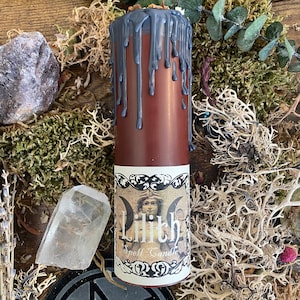 May include: A brown and black spell candle with a label that reads "Lilith Spell Candle". The candle is decorated with a black wax drip and a vintage-style image of a woman with a crown.