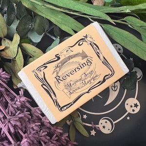 May include: A bar of "Reversing Magical Soap" by Madame Phoenix, wrapped in clear plastic. The label is tan with black decorative elements and text. The soap is surrounded by green leaves and purple lavender.