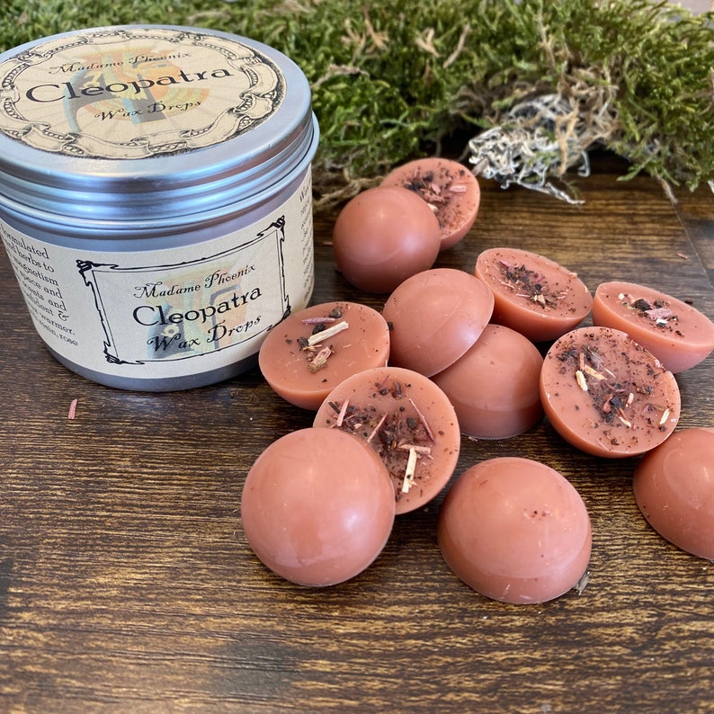 May include: A tin of wax melts labelled "Madame Phoenix Cleopatra Wax Drops". The tin is open and there are several brown wax melts scattered around it. The wax melts have a speckled appearance and some have small pieces of dried herbs on top.