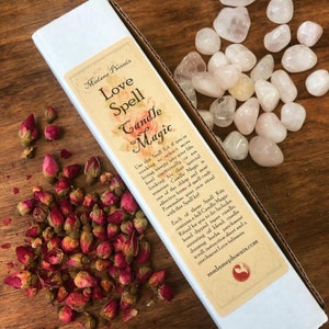 May include: A white box with a pink and white label that reads "Madame Phoenix Love Spell Candle Magic". The label has text describing the contents of the kit, which includes a candle, oil, herbs, and a parchment scroll. The box is sitting on a wooden surface with pink rose buds and pink stones.