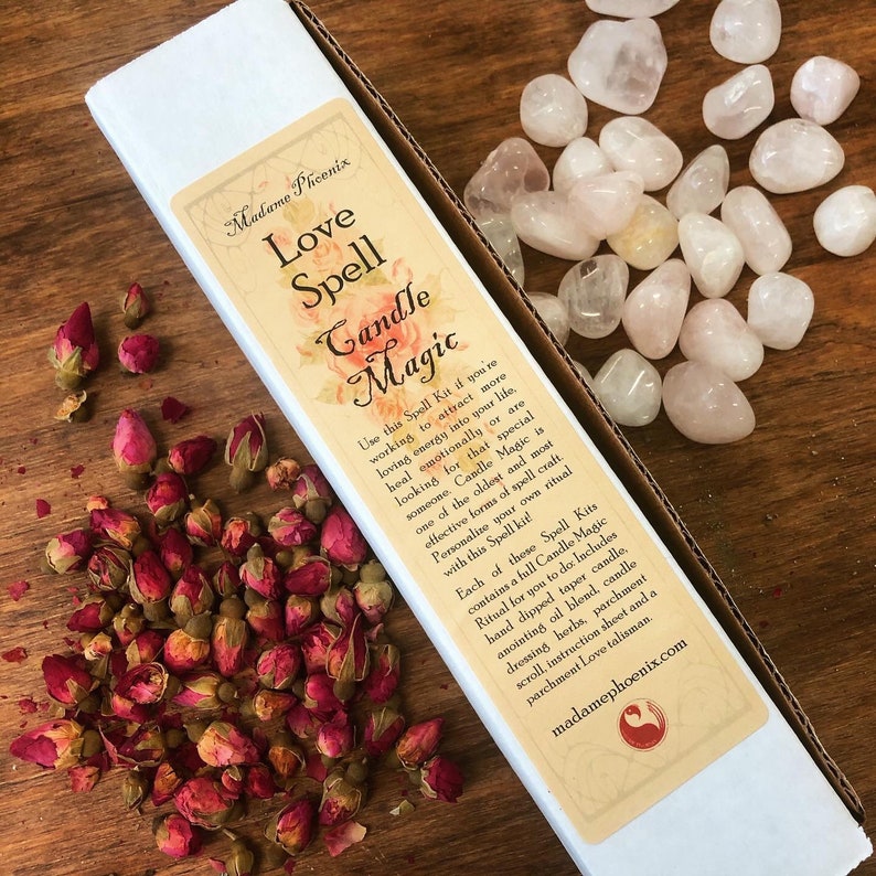 May include: A white box with a pink and white label that reads "Madame Phoenix Love Spell Candle Magic". The label has text describing the contents of the kit, which includes a candle, oil, herbs, and a parchment scroll. The box is sitting on a wooden surface with pink rose buds and pink stones.