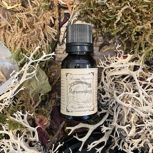 May include: A small brown glass bottle with a black cap and a white label. The label says "Madame Phoenix's Reversing Magical Oil Blend".