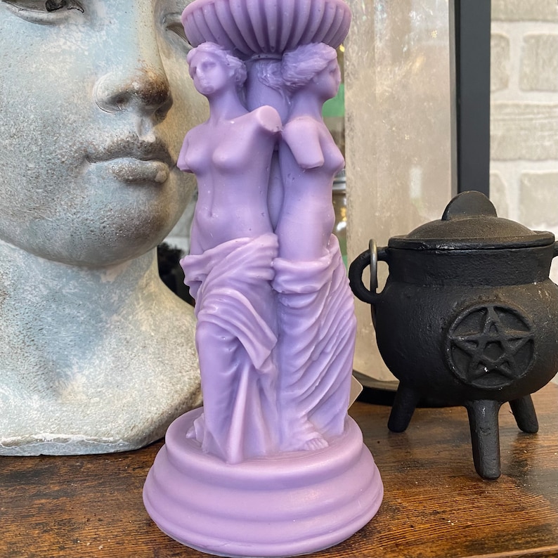 May include: Purple candle holder with two female figures sculpted in a draped style. The candle holder has a round base and a decorative top.