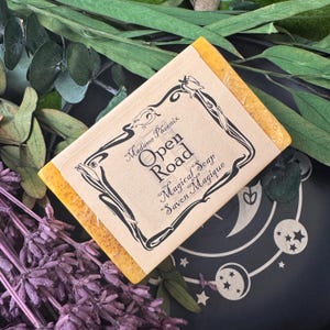 以下が含まれることがあります： 「Open Road Magical Soap Savon Magique」の黒い文字が書かれたベージュ色のラベルが付いた黄色の石鹸。石鹸は緑と紫のラベンダーの小枝に囲まれています。