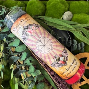May include: A cylindrical spell candle with a label featuring the text "Madame Phoenix Spell Candle / Bougie Magique" and "Love Reversing amour inverse". The label has a pink and cream color scheme with floral and geometric designs.
