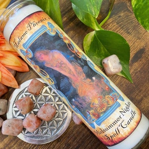 Op de afbeelding: Een witte kaars met een kleurrijk etiket waarop staat "Madame Phoenix's Midsummer Nights Spell Candle". Het etiket toont een vrouw in een vloeiende jurk die in een bosrijke omgeving staat.