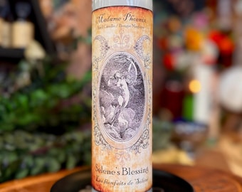 Op de afbeelding: Een hoge cilindrische glazen kaars met lichtgrijze was aan de binnenkant. Het etiket bevat de tekst Madame Phoenix, Spell Candle en Selene's Blessing, met een ovale illustratie van een figuur die naar een maansikkel reikt.
