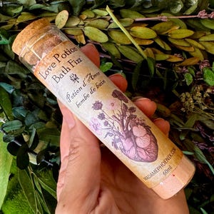 May include: A cylindrical glass vial with a cork stopper containing pink bath fizz. The label reads Love Potion Bath Fizz, Potion d'Amor bombe de bain, and MADAMEPHOENIX.COM, featuring an anatomical heart illustration with flowers and 60ml text.