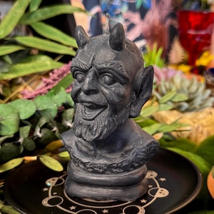 Peut inclure: Une bougie gris foncé en forme de tête de diable avec des cornes, une barbe et une expression souriante. La bougie est posée sur une assiette noire avec un motif céleste. Le fond comprend de la verdure et des décorations colorées.