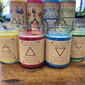 May include: Four glass candle jars with labels that say "Elemental Air Spell Candle", "Elemental Fire Spell Candle", "Elemental Water Spell Candle", and "Elemental Earth Spell Candle". The candles are yellow, red, blue, and green, respectively. Each candle has a different symbol on the label.