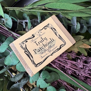 Peut inclure: Un savon "Truly Patchouli Magical Soap" dans un emballage en papier beige avec du texte décoratif noir. Le savon est entouré de feuilles vertes et de fleurs violettes, créant une esthétique naturelle.