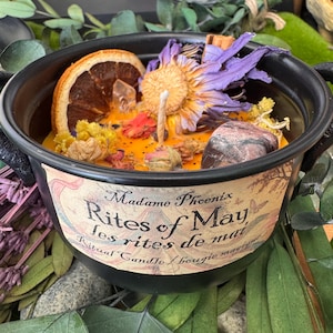 May include: A black cauldron-shaped candle with a label that reads "Madame Phoenix Rites of May". The candle is filled with orange wax and decorated with dried flowers, a crystal, and a stone. The candle has a wick.