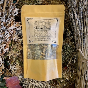 May include: A brown paper pouch with a clear window, containing a blend of dried herbs and flowers. The pouch has a label that reads "Madame Phoenix's Moon Time Regles de Lune Tea Blend Melange de the".
