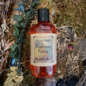May include: A clear orange bottle with a brown label that reads "Madame Phoenix's Pumpkin Patch Bubble Bath". The bottle is sitting on a bed of moss and other natural materials.