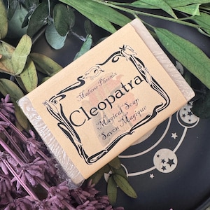 May include: A bar of soap wrapped in clear plastic with a tan label. The label reads "Cleopatra Magical Soap Savon Magique" in black script with decorative floral accents. The soap is surrounded by greenery and purple flowers.