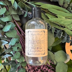 May include: A clear bottle of floor wash with a black cap. The label reads "Madame Phoenix Rosemary & Lime romarin et citron vert Floor Wash Lavage des sols". The bottle is surrounded by green and purple foliage.