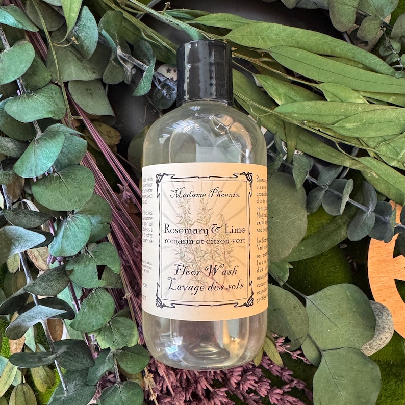 May include: A clear bottle of floor wash with a black cap. The label reads "Madame Phoenix Rosemary & Lime romarin et citron vert Floor Wash Lavage des sols". The bottle is surrounded by green and purple foliage.