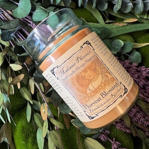 May include: A clear glass jar candle filled with orange wax. The label features the text Madame Phoenix Spell Candle/Bougie Magique and Harvest Blessing B&eacute;n&eacute;diction des R&eacute;coltes, along with an illustration of a person with long hair.