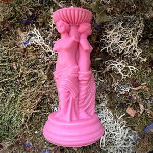 May include: Pink candle holder shaped like two women standing back to back. The candle holder is made of wax and has a detailed design.