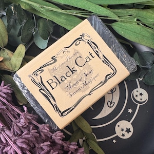 May include: A bar of black soap wrapped in clear plastic, with a tan paper label. The label reads "Black Cat Magical Soap Savon Magique" in black script and decorative borders. The soap is surrounded by dried lavender and greenery.