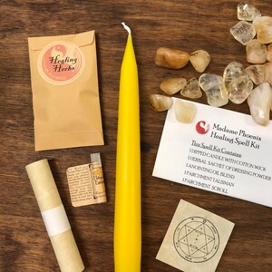 May include: A yellow candle, a small vial of oil, a rolled parchment scroll, a small pouch, and a small card with the text "Madame Phoenix Healing Spell Kit" and a list of the kit's contents. The kit is on a wooden surface with a scattering of clear and brown stones.