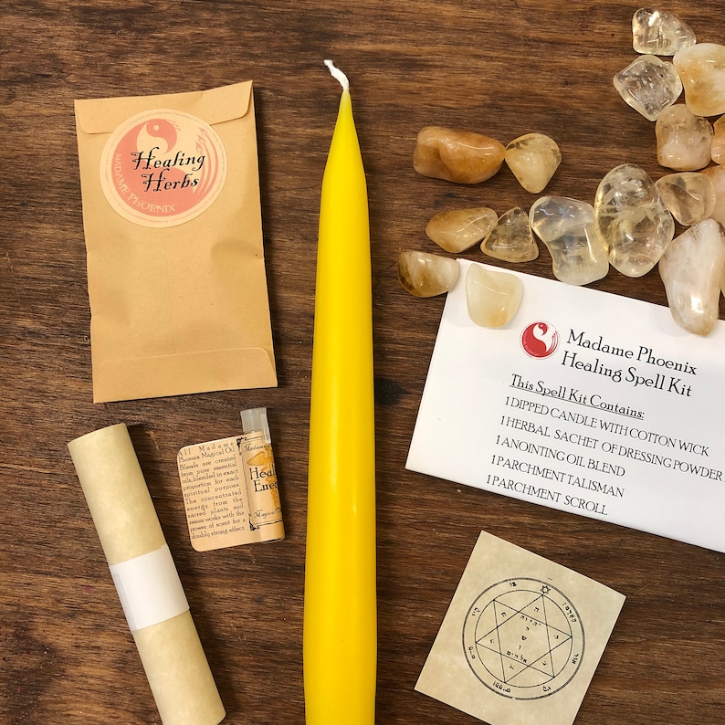 May include: A yellow candle, a small vial of oil, a rolled parchment scroll, a small pouch, and a small card with the text "Madame Phoenix Healing Spell Kit" and a list of the kit's contents. The kit is on a wooden surface with a scattering of clear and brown stones.