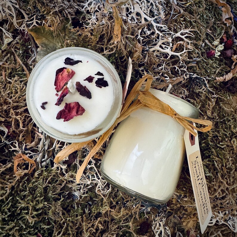 May include: Two soy wax candles, one with dried rose petals on top. The candle on the right has a label that reads "Love Blends Soy Candles, Hand Poured with Love".
