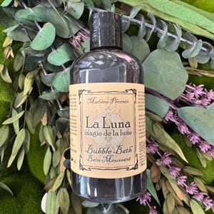 May include: A dark grey bubble bath bottle with a black cap. The label reads "Madame Phoenix La Luna magie de la lune Bubble Bath Bain Moussant". The bottle is surrounded by green and purple foliage.