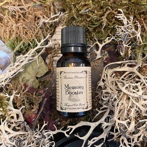 Puede incluir: Una pequeña botella de vidrio marrón con una tapa negra, etiquetada "Madame Phoenix's Memory Booster Magical Oil Blend". La botella está rodeada de musgo seco y otros elementos naturales.