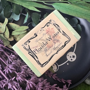May include: A bar of green soap with a decorative label that reads "Florida Water Magical Soap Savon Magique." The label has a floral design and is framed by black decorative lines. The soap is on a black plate with a celestial design.