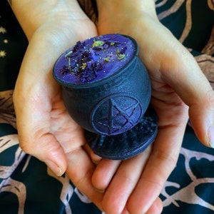 May include: A black cauldron candle holder with a pentagram design. The candle is purple and has dried flowers and glitter on top.