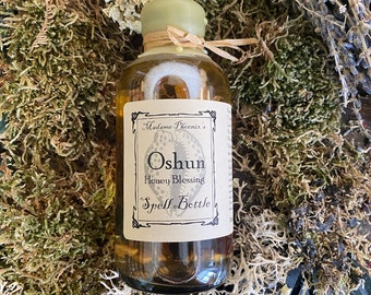 May include: A glass bottle with a brown label that reads "Madame Phoenix's Oshun Honey Blessing Spell Bottle". The bottle is filled with a clear liquid and has a green lid.