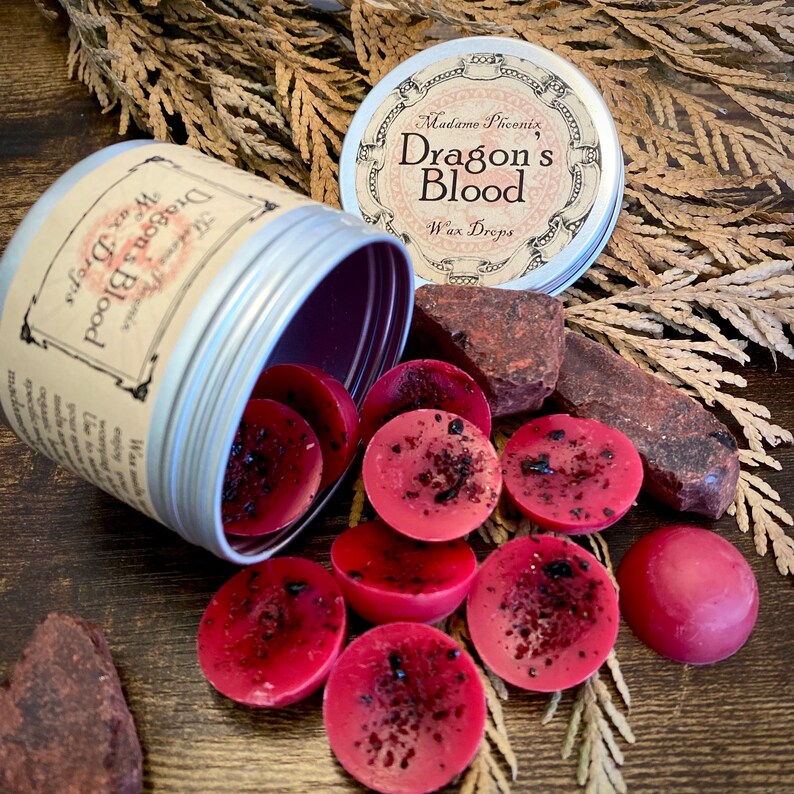 May include: A silver tin with a red and black label that reads "Madame Phoenix, Dragon's Blood Wax Drops". The tin is open and red wax melts are spilling out. The wax melts are round and have a dark red colour with black flecks.