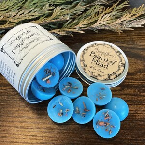 May include: A silver tin with a blue label that reads "Madame Licenix Peace of Mind Wax Drops". The tin is open and blue wax melts are spilling out.
