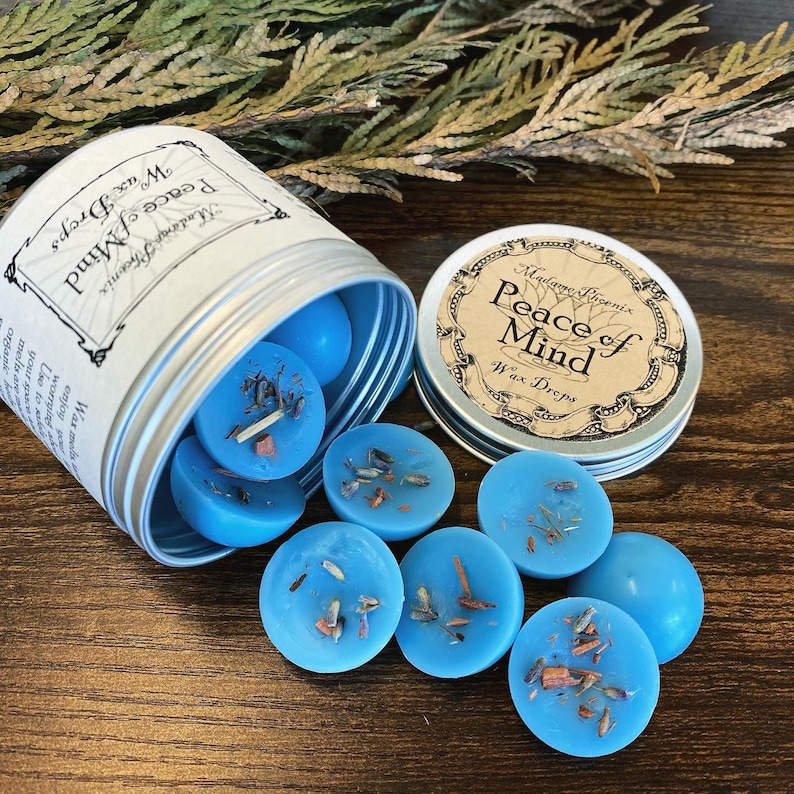 May include: A silver tin with a blue label that reads "Madame Licenix Peace of Mind Wax Drops". The tin is open and blue wax melts are spilling out.