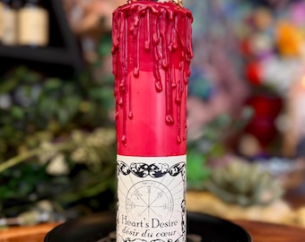 May include: A cylindrical red candle featuring a thick layer of red wax dripping from the top. The base is wrapped in a white label with a black circular emblem and the text Heart's Desire désir du cœur printed in a decorative black frame.