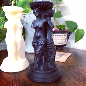 May include: Black candle holder featuring three sculpted figures of women. The figures are standing on a black base with a decorative rim.