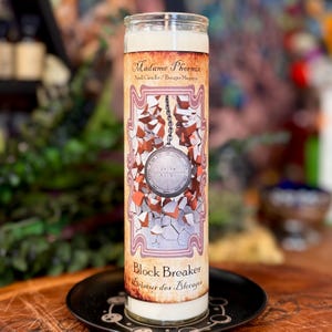 May include: A tall, cylindrical glass candle with a white wax core. The label features the text Madame Phoenix, Spell Candle, Block Breaker, and Briseur des Blocages. The graphic depicts a silver circular seal suspended by a chain over cracked stone.