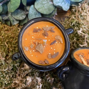 May include: A black cauldron candle with an orange wax scent, filled with cinnamon sticks and gold glitter.