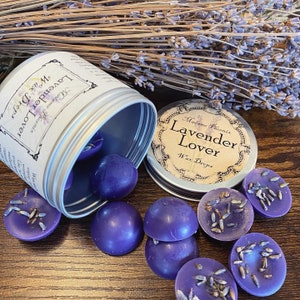 May include: A silver tin with a label that reads "Madame Phoenix Lavender Lover Wax Drops" and a purple wax melt inside. The tin is open and the wax melts are spilling out. The wax melts are round and have a lavender scent.