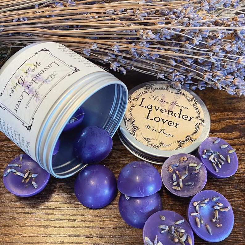 May include: A silver tin with a label that reads "Madame Phoenix Lavender Lover Wax Drops" and a purple wax melt inside. The tin is open and the wax melts are spilling out. The wax melts are round and have a lavender scent.