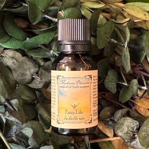 May include: A small amber glass bottle with a black ribbed cap, containing Madame Phoenix magical oil. The label features a sunrise illustration, the text "Easy Life la belle vie," and additional descriptive text in English and French.