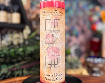May include: A tall cylindrical red wax candle in a clear glass jar. The label features the text Madame Phoenix, Spell Candle, Heart's Desire, and Désir du Cœur, along with a circular geometric sigil design and faint illustrations of pink roses.
